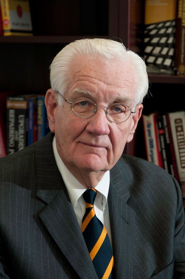 A man in a suit and tie stands in front of a bookshelf