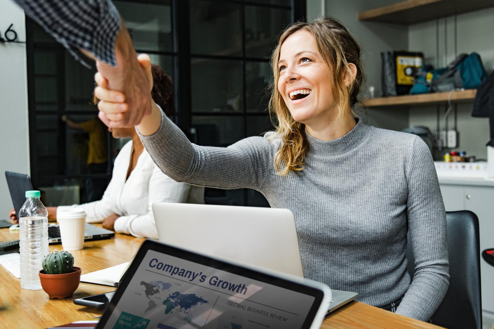 A woman is shaking hands with a man while sitting at a table with a laptop.