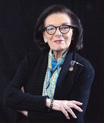 A woman wearing glasses and a scarf is standing with her arms crossed.