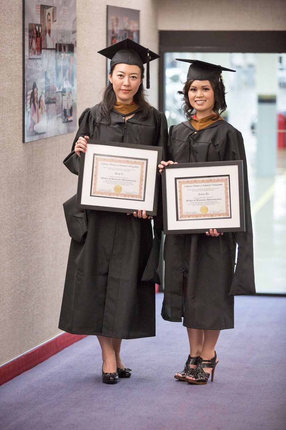 Two female graduates are holding their diplomas in a hallway.