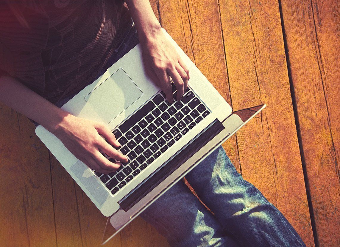 A person is typing on a laptop computer while sitting on a wooden floor.