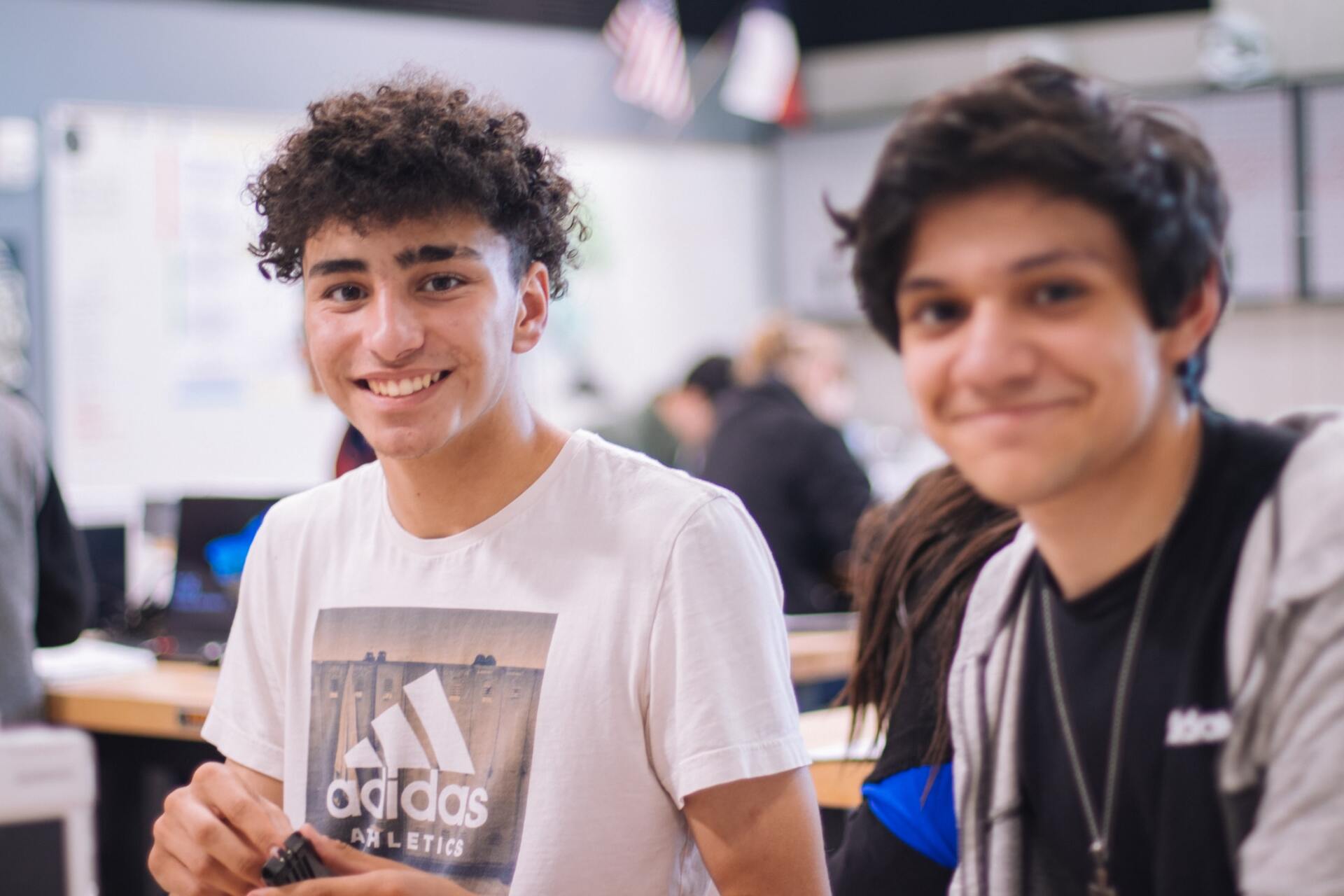 Two young men are sitting next to each other in a classroom and smiling . one of the men is wearing an adidas shirt.
