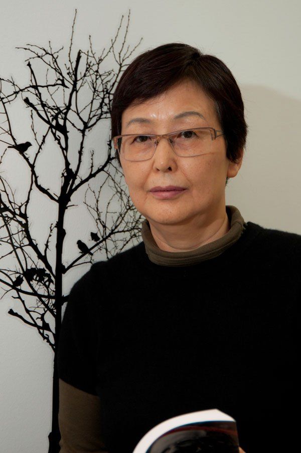 A woman wearing glasses is holding a book in front of a tree with birds on it